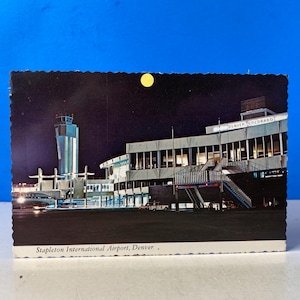 May include: Vintage postcard featuring Stapleton International Airport in Denver, Colorado, at night. The image shows the airport terminal, control tower, and a full moon. The postcard has a scalloped edge.
