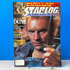 Puede incluir: Una portada de la revista Starlog vintage con Sting como Feyd de la película Dune. La portada incluye el título "Dune" y artículos sobre películas de ciencia ficción. La revista se muestra sobre un soporte blanco sobre un fondo azul.