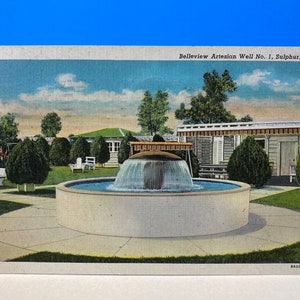 Puede incluir: Una postal que representa una fuente en un entorno de parque. La fuente está hecha de piedra y tiene una cuenca redonda. El agua fluye desde la parte superior de la fuente hasta la cuenca. La fuente está rodeada de hierba verde y árboles. La postal también presenta un edificio en el fondo. El texto en la postal dice "Belleview Artesian Well No. 1, Sulphur, Okla."