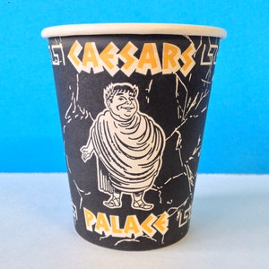 May include: A black paper cup with a white and yellow design. The cup features a cartoon image of a man in a toga and the text "Caesar's Palace".