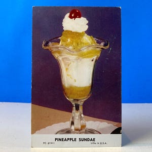 May include: A vintage postcard featuring a pineapple sundae in a clear glass. The sundae has layers of yellow pineapple, white ice cream, and a cherry on top. The postcard is labeled "Pineapple Sundae."
