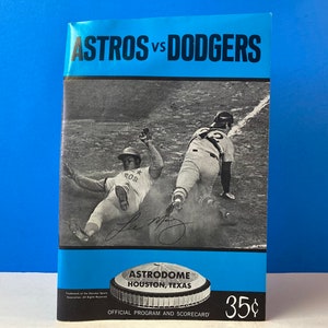 May include: A black and white photo of two baseball players in action. The player on the left is sliding into home plate while the player on the right is trying to tag him out. The photo is from a 1970s baseball game between the Astros and Dodgers. The text on the program reads "ASTROS vs DODGERS ASTRODOME HOUSTON, TEXAS OFFICIAL PROGRAM AND SCOREBOARD 35¢".
