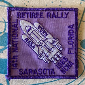 May include: A square, purple embroidered patch with a white NASA space shuttle design. Text includes "RETIREE RALLY", "14th NATIONAL", "SARASOTA", "FLORIDA", and "1985 NCHA". The patch has a purple border.