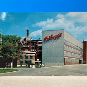May include: A vintage postcard featuring a photo of the Kellogg's factory. The building is a large, multi-story structure with a red sign that reads "Kellogg's". The factory is surrounded by trees and a paved road.