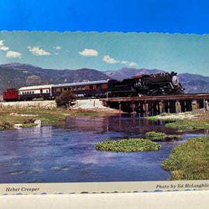 Pode incluir: Postal vintage com um comboio a vapor preto a atravessar uma ponte de madeira sobre um rio. O postal mostra uma paisagem cénica com montanhas ao fundo. O texto "Heber Creeper" e "Photo by Ed McLaughlin" é visível.