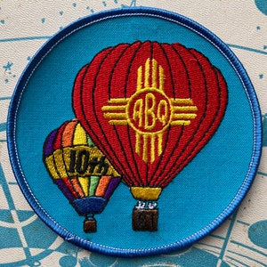 May include: A blue circular patch with a red and yellow hot air balloon with the text "ABQ" and a smaller rainbow hot air balloon with the text "10th" on it.