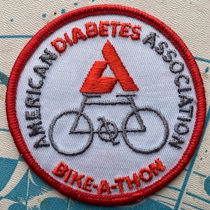 Pode incluir: Um remendo bordado em vermelho, branco e cinza com o texto "American Diabetes Association Bike-A-Thon". O remendo apresenta uma bicicleta estilizada com a letra "A" no topo.