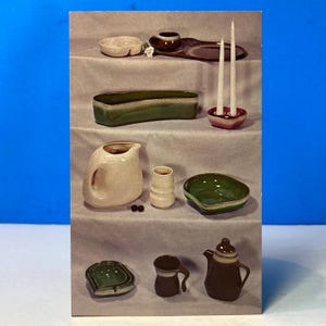May include: A collection of ceramic tableware including a cream pitcher, a green oblong dish, a small pink candle holder with two white taper candles, a green bowl, a brown teapot, a brown mug, and a green ashtray arranged on tiered shelves.