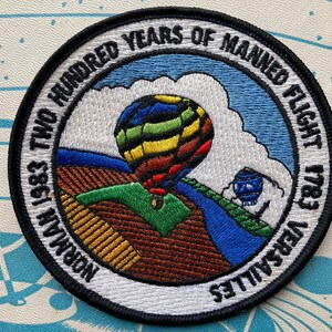 May include: A white circular patch with black trim, featuring a colourful hot air balloon rising above a green and brown landscape. The text "Two Hundred Years of Manned Flight 1783-1983 Versailles" is embroidered around the edge.