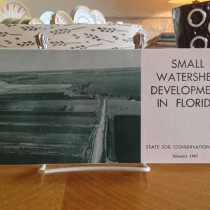 Puede incluir: Una fotografía aérea en blanco y negro de un campo con hileras de cultivos. La imagen forma parte de un folleto titulado "Small Watershed Development in Florida" con el texto "State Soil Conservation Board January 1967".