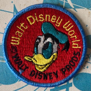 Pode incluir: Um emblema bordado circular com fundo vermelho e borda azul. O centro exibe a cabeça do personagem Donald Duck em branco, preto e amarelo. O texto Walt Disney World está bordado em amarelo, com Walt Disney Prods em preto.