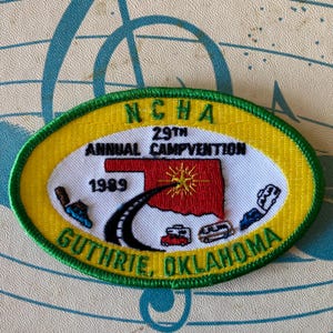 May include: Embroidered patch with a yellow border and a green stitched edge. The patch features the text "NCHA 29th ANNUAL CAMPVENTION 1989 GUTHRIE, OKLAHOMA" and an image of the state of Oklahoma with a road and vehicles.