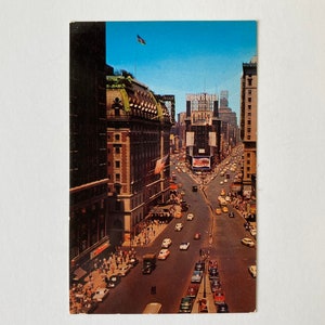Puede incluir: Postal vintage con una vista en ángulo alto de Times Square en la ciudad de Nueva York. La imagen muestra coches clásicos, edificios altos y vallas publicitarias bajo un cielo azul claro. Se ve una bandera americana.