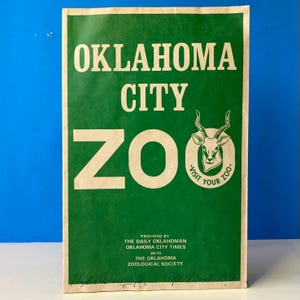 May include: A rectangular paper sign with a green background and cream-colored text reading OKLAHOMA CITY ZOO. It features an illustration of an antelope head and the text VISIT YOUR ZOO, plus credits to The Daily Oklahoman and Zoological Society.