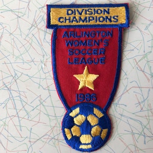 May include: A red, blue, and gold embroidered patch with a soccer ball, a star, and the text "Division Champions Arlington Women's Soccer League 1986".