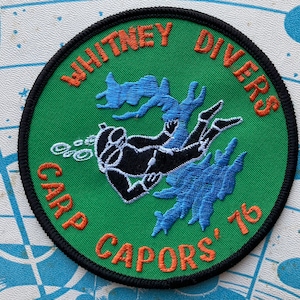 以下が含まれることがあります： 緑色の背景にオレンジ色の糸で「WHITNEY DIVERS CARP CAPORS' 76」と刺繍されたパッチ。黒いダイバーのシルエットと青い水の要素が特徴です。パッチには黒い縁取りがあります。