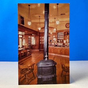 May include: A vintage postcard featuring the interior of a shop with wooden floors and display cases. A dark, ornate stove stands in the foreground, with hanging lamps and a central pillar. The shop appears to sell various items.