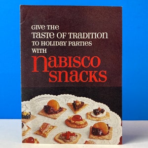 May include: A vintage Nabisco snacks cookbook with a black cover and red text that reads "Give the Taste of Tradition to Holiday Parties with Nabisco Snacks". The cover features a photo of a plate of crackers with various toppings.