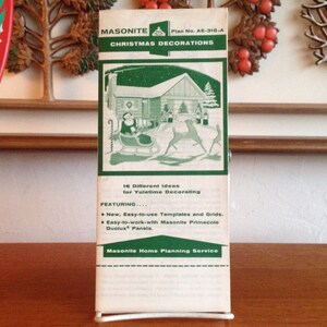 May include: A vintage Masonite Christmas decorations pamphlet with a green and white color scheme. The pamphlet features a small illustration of a house with a Christmas tree inside. The text on the pamphlet reads "Masonite Christmas Decorations" and "Plan No. AE-318-A".