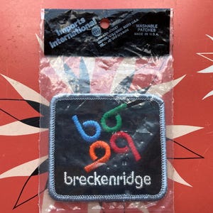 Puede incluir: Un parche bordado cuadrado negro con la palabra "breckenridge" en blanco. El parche presenta números y letras estilizados y coloridos en azul, verde, naranja y rojo. El parche está en un paquete de plástico transparente con el logotipo "Imports International".