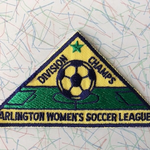 May include: A yellow triangular patch with a blue border and green field. The patch reads "Division Champs" with a soccer ball in the center and a green star at the top. The bottom of the patch reads "Arlington Women's Soccer League".