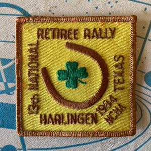 May include: Yellow square patch with brown stitched border and text. The patch reads "RETIREE RALLY" above a brown horseshoe with a green four-leaf clover. Below the horseshoe is "HARLINGEN 1984 NCHA". The left side reads "13th NATIONAL" and the right side reads "TEXAS".