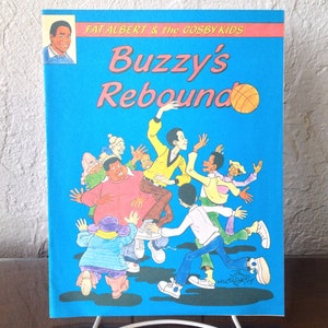 May include: A blue book cover with the title "Buzzy's Rebound" in yellow and red lettering. The cover features a cartoon illustration of a group of children playing basketball, including a basketball with orange and brown stripes.