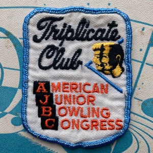 Könnte beinhalten: Gestickter Aufnäher mit dem Text "Triplicate Club" in schwarzer Schrift über "American Junior Bowling Congress" in orangefarbenen Blockbuchstaben. Der Aufnäher hat einen blauen Rand und eine Illustration von zwei Bowlingfiguren.