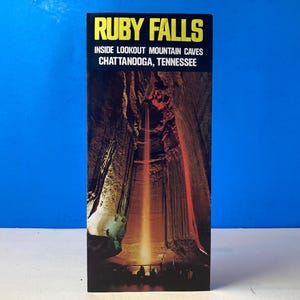 Könnte beinhalten: Eine Vintage-Postkarte für Ruby Falls, Inside Lookout Mountain Caves, Chattanooga, Tennessee. Die Postkarte zeigt ein vertikales Bild der Höhle mit einem Wasserfall, der von einem Lichtstrahl beleuchtet wird. Der Text "RUBY FALLS" ist gelb.