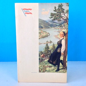 May include: A vintage travel poster featuring a woman in traditional Norwegian clothing standing by a tree overlooking a river and mountains. The poster has the text "N.A.L" and "VALDORES" printed on it.