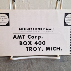 May include: A white business reply mail envelope with black text. The text reads "BUSINESS REPLY MAIL", "FIRST CLASS PERMIT NO. 26 - TROY, MICHIGAN.", "AMT Corp.", "BOX 400", and "TROY, MICH.".