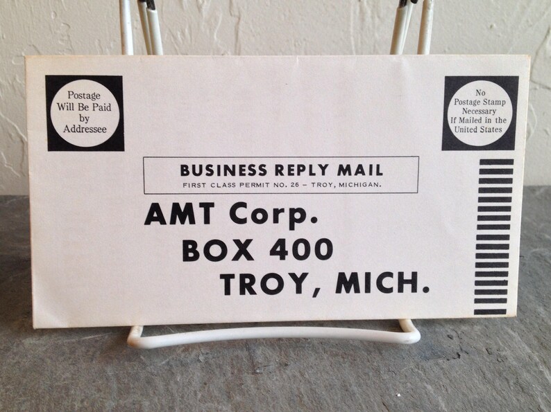 May include: A white business reply mail envelope with black text. The text reads "BUSINESS REPLY MAIL", "FIRST CLASS PERMIT NO. 26 - TROY, MICHIGAN.", "AMT Corp.", "BOX 400", and "TROY, MICH.".