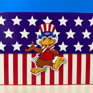 May include: A red, white, and blue eagle mascot wearing a hat with the Olympic rings. The mascot is standing on a red, white, and blue striped background.