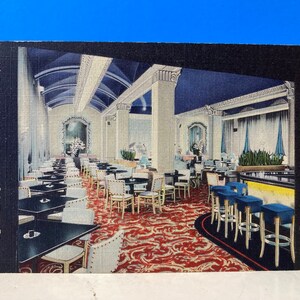 Op de afbeelding: Vintage ansichtkaart met de Cotillion Lounge in het Jung Hotel in New Orleans, LA. Het interieur toont tafels, stoelen en een bar met blauwe krukken. Het tapijt heeft een rood-wit patroon en het plafond is blauw.