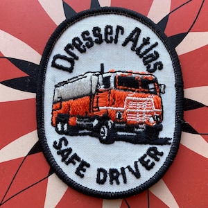 Może przedstawiać: Biała naszywka haftowana z czarną obwódką. Naszywka przedstawia czerwony i biały samochód ciężarowy z tekstem "Dresser Atlas Safe Driver" wokół samochodu.