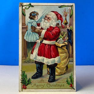 Puede incluir: Postal navideña vintage con Papá Noel sosteniendo a un niño. Papá Noel viste un traje rojo con ribetes blancos y botas negras. Una gran bolsa de juguetes está a su lado. Las palabras "Merry Christmas" están en la parte inferior.