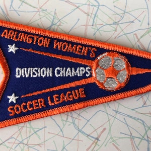 May include: A blue and orange embroidered patch with the text "Arlington Women's Division Champs Soccer League" and a white soccer ball with an orange star in the center.