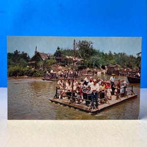 May include: Vintage postcard featuring a river scene with several wooden rafts carrying people. A small building and lush greenery are visible on the riverbank. The postcard has a blue background.