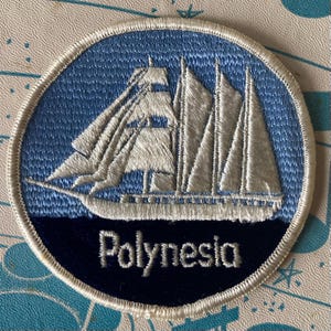 May include: A circular embroidered patch featuring a white sailing ship on a light blue background. The bottom section is a dark navy blue crescent with the word Polynesia embroidered in white text. The entire patch has a white stitched border.
