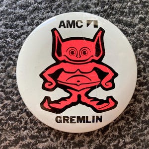 May include: A round, white button featuring a red gremlin cartoon character with black outlines. The button has the text "AMC" at the top and "GREMLIN" at the bottom. The gremlin has large eyes and ears.