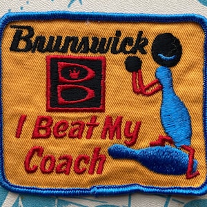 Può includere: Una toppa ricamata gialla e blu con il testo "Brunswick" e "I Beat My Coach". Accanto al testo è raffigurata una figura di un cartone animato di una persona che tiene una palla da bowling.