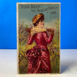 May include: A vintage trade card featuring a person in a pink dress with a bustle and a yellow hat, holding a light-colored parasol. The text reads: STAR SOAP THE BEST FOR FAMILY USE. The background shows green foliage against a blue sky.