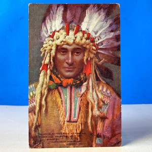 Op de afbeelding: Vintage ansichtkaart met een portret van een Indiaanse man met een gevederde hoofdtooi en traditionele kleding. De afbeelding heeft een sepia-tint met accenten van rood, geel en blauw. Tekst aan de onderkant van de kaart is gedeeltelijk zichtbaar.