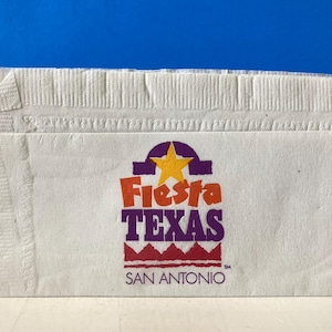 May include: White paper napkin with a purple, yellow, red, and orange logo that reads "Fiesta Texas" and "San Antonio".