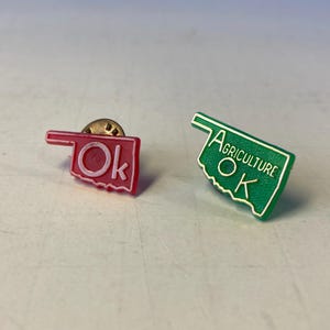 May include: Two Oklahoma state-shaped pins. One is red with "OK" in white lettering. The other is green with "AGRICULTURE OK" in white lettering.
