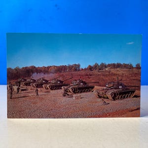 May include: A vintage postcard featuring a military scene with several olive-drab tanks lined up on a gravel surface. Soldiers are present, and the background includes trees and a blue sky. The postcard has a retro aesthetic.