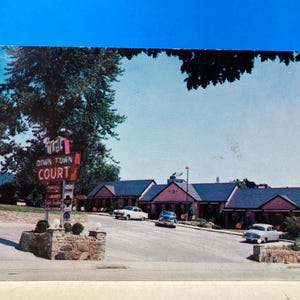 May include: Vintage postcard featuring a motel called "Down Town Court." The image shows a row of motel rooms with cars parked in front. A sign with the motel's name is visible, along with trees and a blue sky.