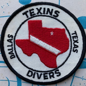 May include: A white and black embroidered patch with a red outline of the state of Texas. The patch reads "Dallas Texans Divers" and has a white scuba diving flag across the state.