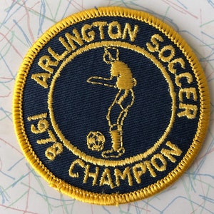 May include: A yellow and blue embroidered patch with the text "Arlington Boys Soccer 1961 Champion". The patch features a stylized image of a soccer player kicking a ball.