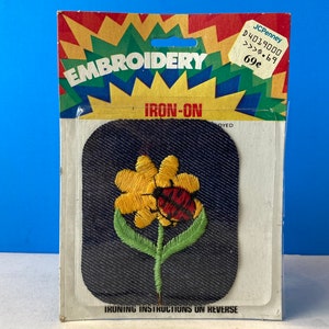 Puede incluir: Parche de bordado termoadhesivo con una flor amarilla con una mariquita roja sobre un fondo azul oscuro. El embalaje incluye el texto "EMBROIDERY IRON-ON" y "IRONING INSTRUCTIONS ON REVERSE".